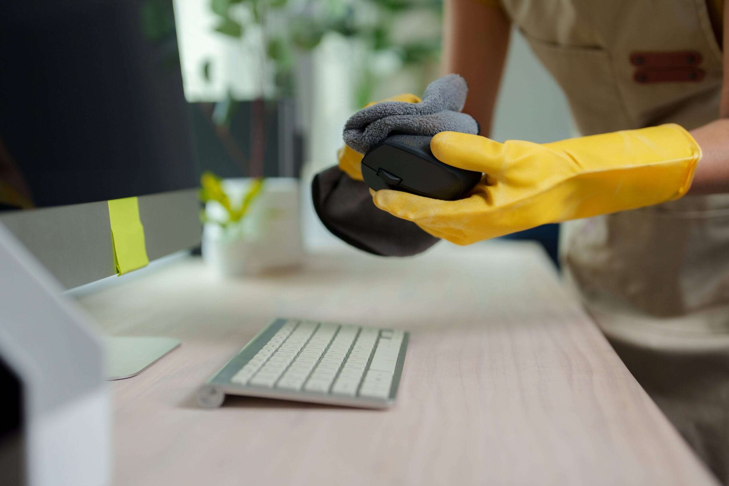 cleaner wiping down a desk in an office, lifting the mouse and wiping it with a yellow microfibre cloth