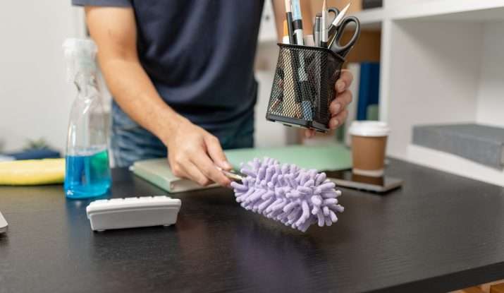 cleaner dusting an office desk, lifting up pen bot and using purple duster in other hand
