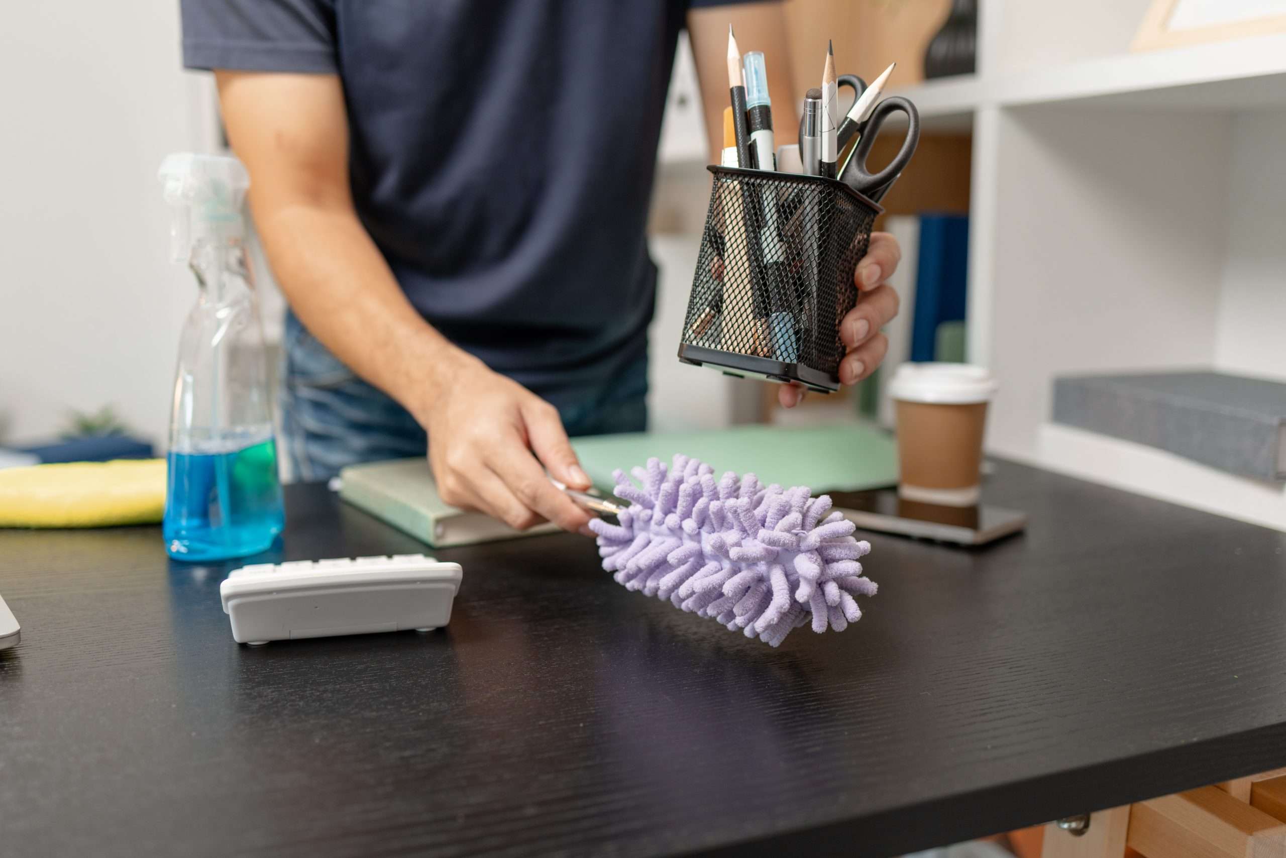 cleaner dusting an office desk, lifting up pen bot and using purple duster in other hand