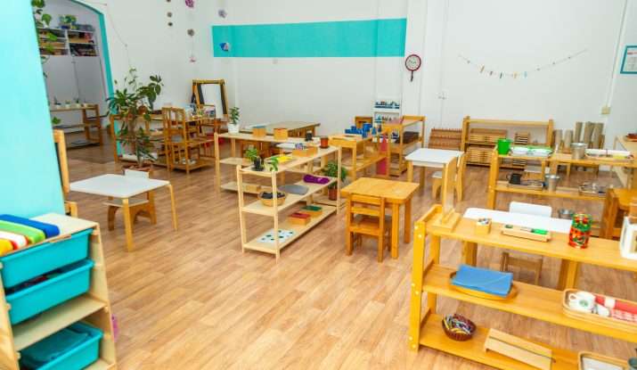 A colorful clean nursery classroom with wooden furniture and educational materials. Plants and decorations create a welcoming environment.